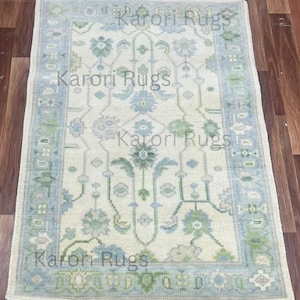 May include: Rectangular rug with a cream-colored field and a border in shades of blue and green. The rug features a floral pattern with stylized flowers and geometric shapes. The text "Karori Rugs" is repeated across the rug.