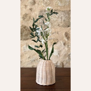 May include: A white wicker vase with a round, bell-shaped design, filled with white flowers and green leaves.