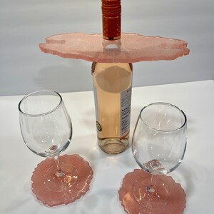 May include: Pink resin wine bottle holder with two matching coasters. The holder is shaped like a cloud and has a hole in the center to hold a bottle of wine. The coasters are round and have a scalloped edge.