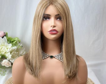 May include: A blonde wig with a blunt cut, styled on a mannequin head. The wig has a natural-looking color and texture. The mannequin is displayed on a woven mat, with a decorative necklace. The background is a soft white.