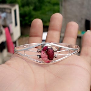 Pode incluir: Uma pulseira de punho prateada com uma pedra preciosa oval composta em tons de rosa e bordô. A pulseira tem um design aberto com elementos decorativos de filigrana e pequenas contas prateadas que acentuam a pedra.
