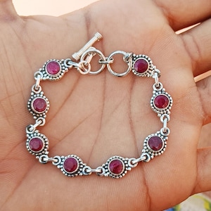 Op de afbeelding: Een zilveren armband met een schuifgesp heeft een ketting met zeven ronde rode edelstenen die in zilver zijn gezet.