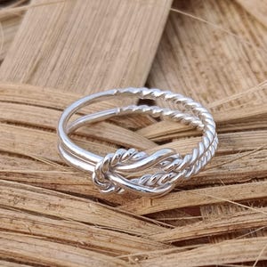 Puede incluir: Un anillo de plata con un diseño de cuerda trenzada y un nudo en el centro.