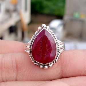 May include: A silver ring featuring a large, pear-shaped, deep red gemstone. The gemstone is surrounded by a twisted rope-like border and small silver beads. The ring's band is a simple, split design.