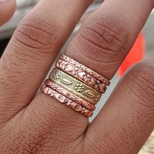 Puede incluir: Un conjunto de cinco anillos apilables hechos de cobre y latón. Los anillos tienen diferentes patrones y texturas, incluyendo corazones, hojas y diseños geométricos.