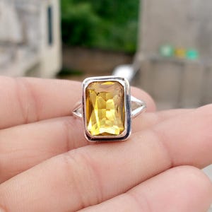 Może przedstawiać: Srebrny pierścionek z dużym, prostokątnym, żółtym kamieniem szlachetnym. Kamień jest osadzony w prostokątnym obramowaniu, a pierścionek ma prosty, elegancki design. Pierścionek jest pokazany na rozmytym tle.
