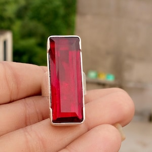May include: A rectangular, red gemstone ring with a silver band. The gemstone is a deep, vibrant red color and is set in a simple, elegant silver setting. The ring is a statement piece, perfect for adding a pop of color to any outfit.