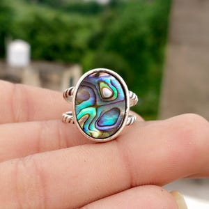 Può includere: Anello in argento con conchiglia di abalone ovale dai riflessi iridescenti blu, verdi e viola. L'anello ha un design a doppia fascia con un dettaglio a corda ritorta. La conchiglia misura circa 2 cm di lunghezza.