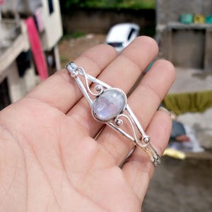 Pode incluir: Uma pulseira de prata com uma pedra da lua oval e iridescente. A pulseira tem um design delicado e vazado com linhas curvas e pequenos detalhes redondos. A pedra da lua é definida em uma moldura. Uma joia.