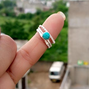Peut inclure: Un ensemble de trois bagues en argent. L'une présente un motif torsadé, une autre est une simple bande, et la troisième est ornée d'une pierre turquoise ronde. Les bagues sont empilées, mettant en valeur un style de bijoux minimaliste.