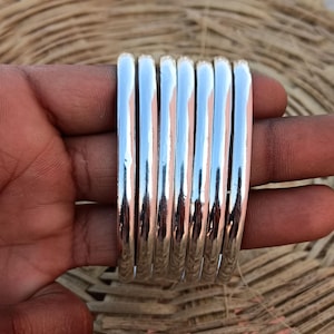 Op de afbeelding: Een set van acht zilveren armbanden, gestapeld en vastgehouden in een hand. De armbanden zijn glad en glanzend en reflecteren licht.