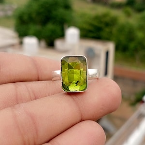 May include: A silver ring featuring a large, rectangular, faceted lime green gemstone set in a simple metal band. The stone displays internal clarity with visible natural inclusions and a bright, translucent, vibrant green color profile.