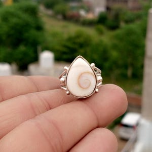 May include: A silver ring featuring a teardrop-shaped shiva eye shell as the centrepiece. The shell is white with a natural orange spiral pattern. The metal band has a polished finish with small decorative silver spheres on each side of the stone.
