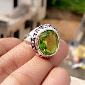 May include: Silver ring featuring an oval, light green gemstone. The ring has an ornate, engraved design around the gemstone. The gemstone is a vibrant, translucent green colour. The ring is held in a hand.