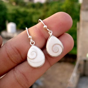 Puede incluir: Pendientes de plata con un colgante blanco en forma de lágrima. Cada colgante presenta un diseño en espiral y está enmarcado por un borde plateado. Los pendientes cuelgan de un gancho plateado.