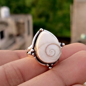 Op de afbeelding: Zilveren ring met een druppelvormige, crèmekleurige steen met een spiraalpatroon. De ring heeft een gedetailleerde zilveren zetting met kleine, decoratieve accenten. De ring wordt op een hand getoond, met een vage achtergrond.