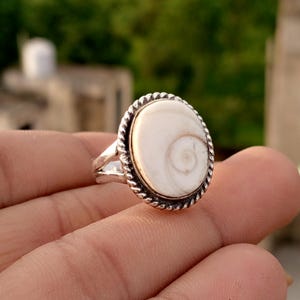 Könnte beinhalten: Silberfarbener Ring mit einem ovalen, cremefarbenen Stein mit Spiralmuster. Der Stein ist von einem gedrehten, seilartigen Rand umgeben. Der Ring ist ein Schmuckstück.