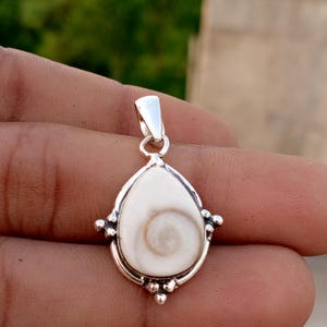 May include: A teardrop-shaped pendant with a white, swirled center and a silver-colored metal frame. The pendant has small, decorative silver accents and a loop for a chain. The background is blurred.