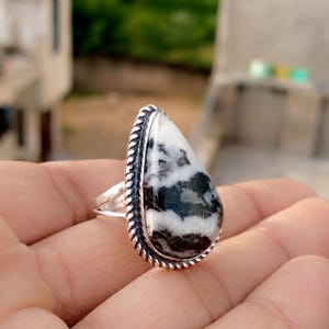 May include: A silver ring featuring a large, teardrop-shaped stone with a black and white zebra jasper pattern. The stone is framed by a twisted rope-like border. The ring is held in a hand, showcasing the jewelry.