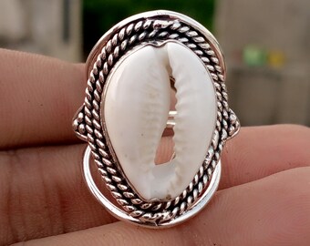 May include: A silver-toned ring featuring a large, oval, white seashell centerpiece. The shell is set within a decorative, textured silver frame with a braided rope design around the shell. The ring has a unique, natural aesthetic.