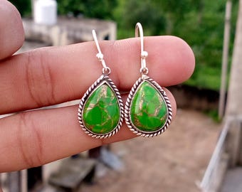 May include: A pair of teardrop earrings featuring a vibrant green stone with gold inclusions, set in silver-coloured metal. The earrings have a rope-style border and a simple hook. The background is blurred, suggesting an outdoor setting. The earrings are a piece of jewellery.
