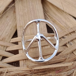 May include: A silver peace sign ring. The ring features the iconic peace symbol, a circle with three lines inside. The ring is set against a woven, light brown background.