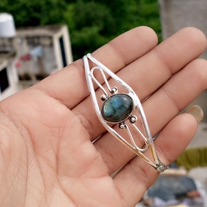Può includere: Un bracciale in argento con una pietra ovale di labradorite iridescente. Il bracciale presenta un design traforato con piccoli dettagli a sfera d'argento. La labradorite mostra sfumature di blu, verde e grigio, creando un effetto visivo accattivante.