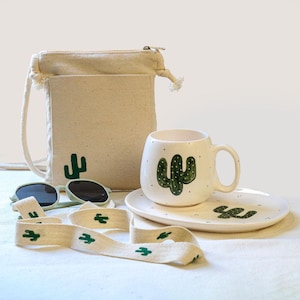Könnte beinhalten: Eine weiße Keramiktasse und eine passende Untertasse mit einem grünen Kaktus-Design. Die Tasse hat einen Henkel und die Untertasse ist oval geformt. Eine weiße Stofftasche mit einem grünen Kaktus-Design und einem passenden grün-weißen Band ist ebenfalls enthalten.