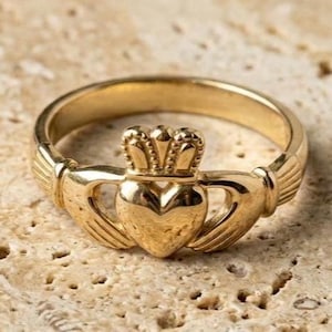 May include: A gold Claddagh ring featuring a central heart topped with a crown, held by two hands on either side of the band. The ring has a polished metallic finish and a traditional design with detailed engravings on the cuffs and crown elements.