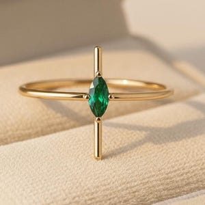 May include: A gold ring with a vertical bar intersecting a horizontal band, featuring a marquise-cut emerald gemstone. The ring is set in a gold band and is displayed in a beige ring box.