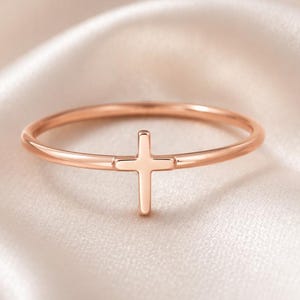 Puede incluir: Un anillo de oro rosa con un diseño de cruz minimalista. El anillo tiene una banda delgada y una pequeña cruz centrada. El anillo se muestra sobre una tela de satén blanca y suave.