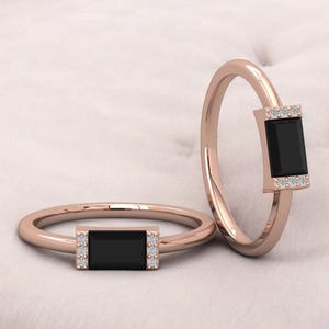 May include: Two rose gold rings, each featuring a rectangular black stone and small white stones. The rings are set against a white, textured backdrop. The design is minimalist.