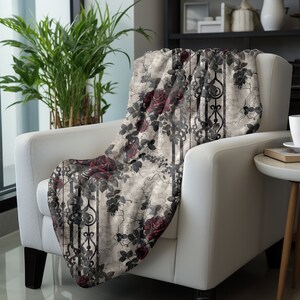 May include: A soft throw blanket featuring a pattern of deep red roses and dark, intricate wrought iron gate designs set against a distressed, light beige background. The fabric displays a repeating motif of climbing rose vines and vertical bars.