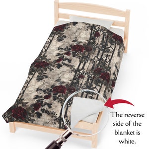 May include: A blanket featuring a pattern of dark red roses and black wrought iron gate designs on a textured beige background. A magnifying glass highlights a corner of the blanket, revealing a plain white reverse side with the text: The reverse side of the blanket is white.