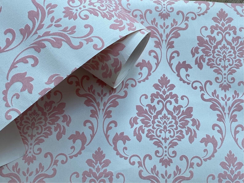 Puede incluir: Rollo de papel tapiz con estampado damasco rosa. El papel tapiz tiene un fondo blanco con un dise&ntilde;o floral rosa.