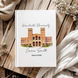 May include: A white hardcover book featuring a watercolor illustration of a brick university building. The text reads Heartville University, Emma Smith, and Class of 2030. The cover design includes a central architectural image with black lettering.