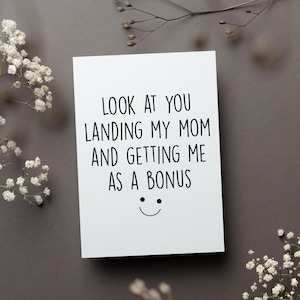 May include: A white greeting card with black text that reads "LOOK AT YOU LANDING MY MOM AND GETTING ME AS A BONUS" with a smiley face. The card is on a gray surface, decorated with white flowers and twigs.