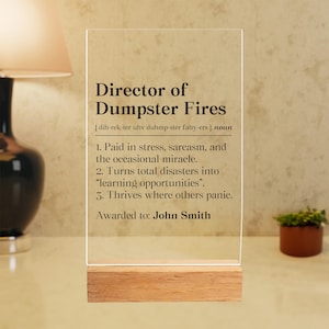 May include: A clear acrylic plaque with the text "Director of Dumpster Fires" and a definition. The plaque is mounted on a wooden base and includes the name "John Smith". The text is in black, and the plaque is displayed on a table.