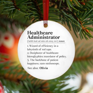 May include: A round white ceramic ornament featuring black text that defines a Healthcare Administrator as a wizard of efficiency, decipherer of healthcare hieroglyphics, and backbone of patient happiness, with the name Olivia listed below.