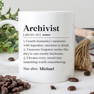 May include: White ceramic mug with the definition of "Archivist" printed in black. The definition includes the word's pronunciation and three descriptive points. Coffee beans are scattered around the mug.