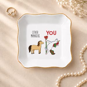 May include: A square white ceramic trinket dish with a gold-trimmed scalloped edge. The center features a brown horse labeled OTHER MANAGERS and a white unicorn with a red and green mane labeled YOU, dancing on a pole with a red bow.