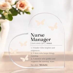 May include: Clear heart-shaped acrylic plaque with the text "Nurse Manager" and a definition. The plaque includes butterfly graphics and the name "Michelle". A vase of flowers is in the background.