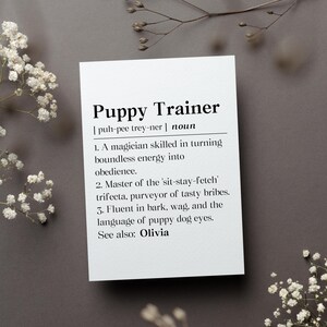 May include: A white rectangular card featuring black printed text that defines the term Puppy Trainer. The text includes a phonetic pronunciation, the noun classification, three numbered definitions, and a See also reference to Olivia.