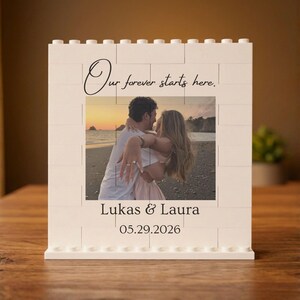 May include: A white rectangular building block frame featuring a central photograph of a couple on a beach at sunset. The text Our forever starts here. is printed above the photo, with Lukas & Laura and 05.29.2026 printed below the image.