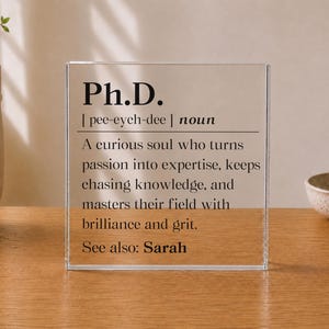 May include: A clear rectangular acrylic block featuring black printed text that defines Ph.D. as a noun, describing a curious soul who turns passion into expertise, keeps chasing knowledge, and masters their field with brilliance and grit. See also: Sarah.
