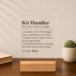 May include: A clear rectangular acrylic plaque standing on a light wood base. The plaque features black printed text defining K9 Handler with three numbered points and the name Ryan at the bottom. The design mimics a dictionary entry style.