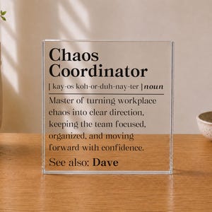 May include: A clear acrylic square plaque featuring black printed text that reads Chaos Coordinator, followed by a phonetic pronunciation, the noun classification, a definition about managing workplace chaos, and the text See also: Dave.