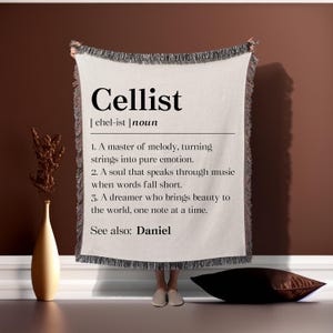 May include: A white throw blanket with fringe edges features a black and white definition of the word "Cellist". The definition reads: "A master of melody, turning strings into pure emotion. A soul that speaks through music when words fall short. A dreamer who brings beauty to the world, one note at a time. See also: Daniel."