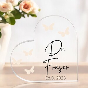 May include: A clear glass heart-shaped plaque with the text "Dr. Fraser Ed.D. 2023" and a design of butterflies.