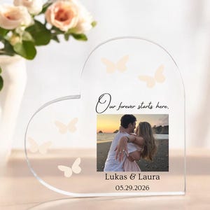 May include: A heart-shaped clear acrylic plaque featuring a printed photograph of a couple on a beach at sunset. The text reads Our forever starts here. Lukas & Laura 05.29.2026. The plaque is decorated with several light-colored butterfly silhouettes.